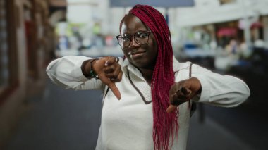 Şehir caddesinde gözlüklü bir kadın, açık havada başparmağını kaldırıyor. Gündüz kent ortamını ön plana çıkarıyor..