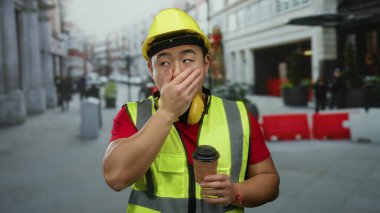 İnşaat işçisi, elinde sert şapka ve güvenlik yeleği olan bir kahveyle şehir sokaklarında esniyor..
