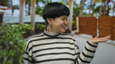 Çizgili kazaklı genç bir kadın açık havada gülümsüyor. Elleri açık, sakin bir bahçe ortamında güvenle poz veriyor..