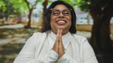 Parkta geniş bir şekilde gülümseyen bir kadın ağaçlarla çevrili, gözlük ve beyaz gömlek takan, mutluluk ve minnettarlık ifade eden, barışçıl neşeli atmosferi vurgulayan bir kadın..