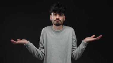 Siyah bir arkaplana karşı kafa karışıklığı içinde omuz silken, gri bir kazak giyen, kararsızlık ve belirsizlik gösteren genç bir adam yakın plan bir portrede.