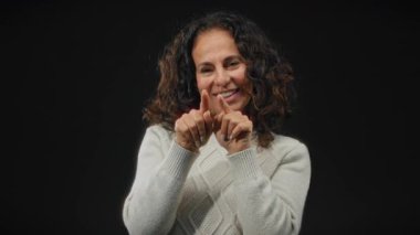 Kıvırcık saçlı, gülümseyen ve işaret eden bir kadın, siyah arka planda soyutlanmış, beyaz bir kazak giyiyor, pozitifliği ve umudu simgeliyor..