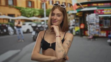Siyah bikinili bir kadın, şehir sokağında gülümsüyor, kafasında güneş gözlüğü var ve şehir binaları ve dükkanlarının geçmişi bulanık..