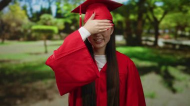 Kırmızı mezuniyet elbisesi giymiş Asyalı genç bir kadın yemyeşil bir parkta tek eliyle neşeli ve kutlama havası yayıyor..