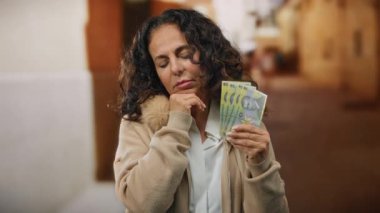 Orta yaşlı bir kadın elinde Romen lei banknotları tutuyor, bir şehir caddesinde düşünceli bir şekilde duruyor, bir şehir ortamında finans ve tefekkür temaları öneriyor..