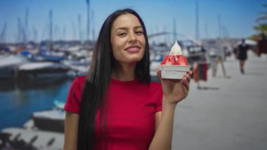 Kırmızı tişörtlü İspanyol kadın güneşli bir sahil limanında teknelerin yanında neşe içinde çilekli dondurma tutuyor..