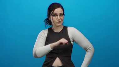 Yumruğunu kameraya doğru uzatan, parlak mavi arka plana karşı kararlı bir ifadeye sahip, gözlük takan ve günlük giyinen, güçlü, kendine güvenen, odaklanmış, güzel bir kadın.