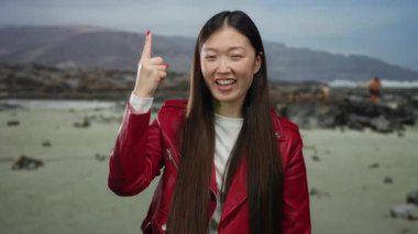 Sahilde kırmızı ceketli, arka planda dağlarla gülümseyen neşeli ifadeler sergileyen ve açık gökyüzünün altında işaret eden bir kadın..