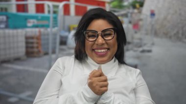 Bir inşaat alanında gülümseyen, iri yarı, gözlüklü, beyaz gömlekli, sanayileşmiş bir ortama güveni ve güveni olan genç, Latin bir kadın..