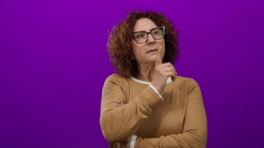 Elini çenesine koyup parlak mor arka plan üzerinde düşünen kahverengi kazak, kıvırcık saç, gözlük takan, düşünceli bir ifade takınan orta yaşlı İspanyol görünümlü bir kadın..