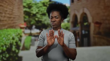 Afro-Amerikalı bir kadın yeşil bir caddede reddedici bir jest yapıyor. Onaylamadığını belirtiyor, jestleri vurguluyor ve açık havada canlı bir şekilde....