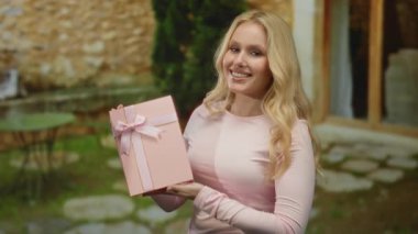 Açık hava restoranında elinde pembe bir hediye tutan genç bir kadın masaları ve taş yolları olan, neşeyle gülümseyen bir kadın..