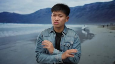 Sahilde genç bir adam kot ceketli parmaklarıyla okyanusa ve arka planda dağlara doğru hareket ediyor.