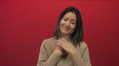 Elleri göğsünde gülümseyen genç bir kadın canlı kırmızı bir arkaplanda mutluluk ve minnettarlığı ifade ediyor..