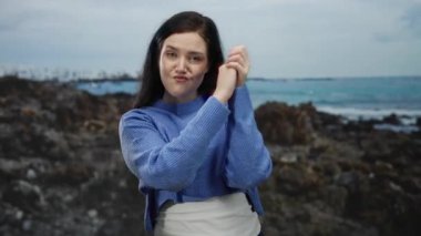 Sahilden hoşlanan bir kadın, okyanus zeminine karşı neşeli bir ifadeyle mavi bir kazak giyiyor ve neşeli özgüveniyle kendini işaret ediyor..