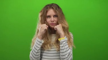 Bir stüdyo sahnesini yumruklarken kadın yumruğunu kaldırıyor. Sarışın, genç, çizgili bir gömlek giymiş, bol saçlı ve kararlı bakışlı bir kadın..