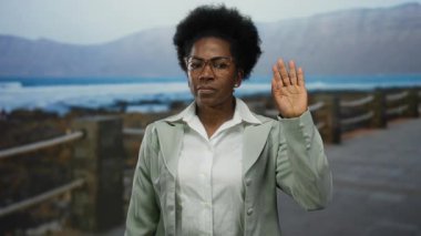 Okyanus manzaralı deniz kenarında el sallayan, gözlüklü ve takım elbiseli, selamlayan, Afro-Amerikan, plaj manzaralı, dingin ve mutlu bir ortam sunan bir kadın..