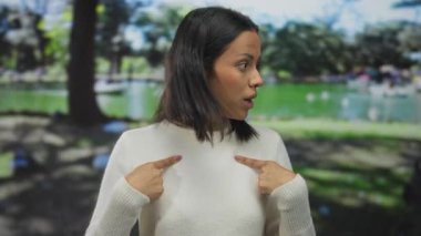 Beyaz kazaklı genç bir kadın, kendini ifade eden bir bakışla, gür yeşil bir parkta, bulanık ağaçlar ve gölde duruyor..