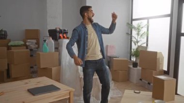 Genç adam, yeni döşenmiş oturma odasındaki kutular arasında neşeyle dans ediyor, eşyalarla dolu modern dairesinde heyecan ve mutluluğu ifade ediyor..