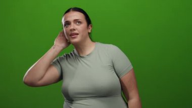 Eli kulağında canlı yeşil bir arka plan karşısında dikkatle dinleyen genç bir kadın merak ve iletişimi işaret ediyor..