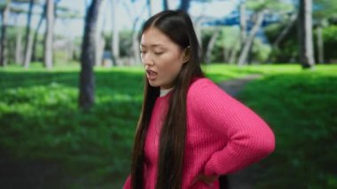 Pembe kazak giyen uzun saçlı kadın arka planda ağaçlarla dolu yemyeşil bir parkta duruyor. Rahatsızlığını dile getiriyor ve açık hava ortamının altında onu geride tutuyor..