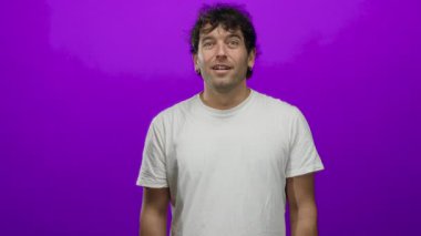 İspanyol bir adam, geniş bir gülümseme ile sevinç ifade ve canlı mor bir arka plana karşı ellerini sımsıkı tutarak gündelik beyaz gömlek giyerek.