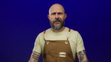 Sakallı, dövmeli, eldiven ve tulumlu bir adam izole edilmiş mavi bir arka plandan endişeli görünüyor.