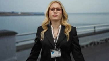 Gözlüklü, sarı saçlı, kumsalda gezinen, siyah takım elbiseli, arka planda okyanus manzaralı bir kadın, profesyonel bir senaryoyu çağrıştırıyor..