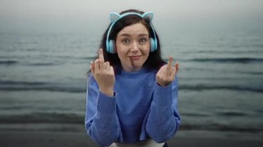 Kulaklık takan ve gülümseyen bir kadın bulutlu bir günde arka planda okyanusla el kol gezerken neşeli ve kaygısız bir anı yakalıyor..