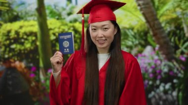 Mezuniyet elbisesi giymiş bir kadın, elinde Kore pasaportuyla, ağaçların arasından süzülen güneş ışığıyla dolu bir parkta kültürel ve eğitimsel başarının bir karışımını sergiliyor.