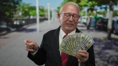 Takım elbiseli kıdemli bir adam güneşli bir sokakta bize dolar tutarken gülümsüyor ve el sallıyor, zenginliği ve güveni ön plana çıkarıyor..