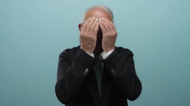 Mavi arka planda kıdemli bir adam. Elleriyle yüzünü kapatıyor. Bu da izole edilmiş profesyonel ortamda neşe, güven ve pozitifliği gösteriyor..
