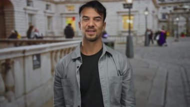 Genç adam dışarıda hareketli bir şehir caddesinde neşeli tavşan kulağı hareketi yapıyor. Bulanık binalar ve arka planda insanlar var. Neşeli ifadesini ve kentsel çevresini vurguluyor..