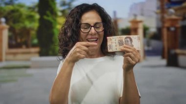 Orta yaşlı İspanyol kadın Meksika pezosu tutuyor, gülümsüyor ve gözlük takıyor, kentsel bir ortamda finansal başarı ve mutluluğu vurguluyor..