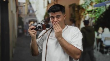 Arka planda bulanık bir kamera tutan genç adam gündüz vakti açık havada beyaz gömleğiyle oyun oynuyor..