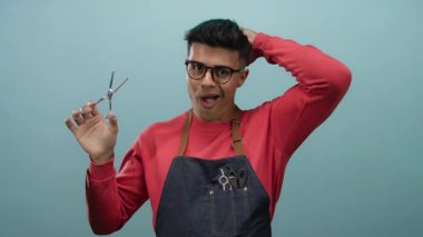 Gözlük ve önlük takan genç adam, kuaför ya da kuaför olarak yeteneklerini gösteren izole edilmiş mavi duvar arkaplanına karşı makas ve makas tutuyor.