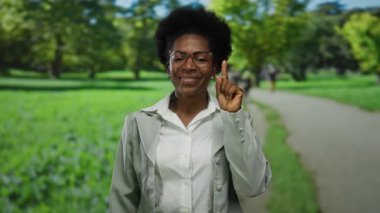 Açık hava parkı ortamında gülümseyen, gözlük takan ve yukarıyı gösteren, yemyeşil ağaçlarla çevrili, berrak gökyüzü manzaralı, pozitiflik ve rahatlık taşıyan bir kadın..