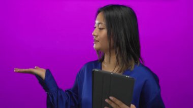 Çinli bir kadın pembe duvarın önünde elinde tablet tutarak mavi bluz giymiş, gülümseyen, genç bir yüz ifadesiyle canlı bir arkaplan karşısında izole bir şekilde jest yapıyor..