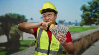 Güvenlik yeleği ve miğfer takan genç bir adam parkta başparmağını sallarken dolar tutuyor..