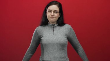 Gözlüklü ve gri kazaklı bir kadın canlı kırmızı arka planda duruyor, yansıtıcı görünüyor, elleri göğsünde, duygu ve tefekkür taşıyor..