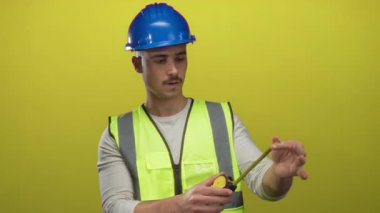 Yansıtıcı yelek giyen genç İspanyol bir adam ve izole edilmiş sarı arka plan duvarına bantlı kasklı ölçüler inşaat işi öneriyor..
