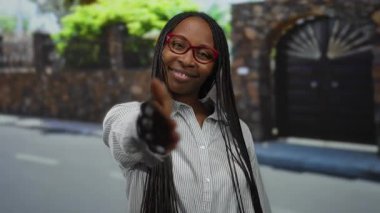 Kıvırcık saçlı, gülümseyen bir kadın, şehir sokaklarında gözlük ve çizgili tişört giyerek el sıkışırken, açık bir şehir ortamında Afrika kökenli Amerikalıların dostça etkileşimini yakalıyor..