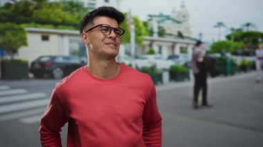 Kırmızı kazaklı genç adam şehir caddesinde özgüvenle gülümsüyor yemyeşil ve modern mimarisi arka planda bulanık..