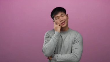 Akıllı telefondan konuşan genç bir adam pembe bir arka plana karşı içerik ifadesiyle gri bir kazak giyiyor ve kapalı bir ortamda sakin bir hava yayıyor..