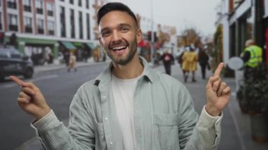 Şehir sokaklarında gülümseyen genç bir adam binalar ve arka planda insanlar, iki eliyle işaret eden, günlük gömlekler giyen, gün ışığında neşe ifade eden
