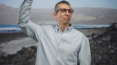Sahilde, güneşli bir havada, açık deniz arka planına karşı, baş parmağıyla jest yapan, gözlük takan ve sıradan gömlekler giyen bir adam..