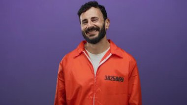 Turuncu hapishane tulumu giymiş İspanyol bir adam canlı mor bir arka plana güvenerek gülümsüyor..