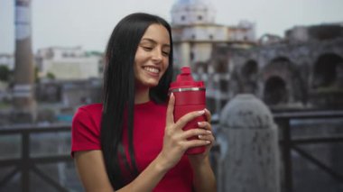 Roma harabelerinde kırmızı bir şişeyle gülümseyen bir kadın, canlı bir ortam ve tarihi mimari sergiliyor, açık havada gençliği ve kültürel keşifleri yansıtıyor..