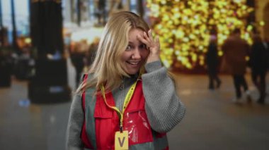 Sarışın bir kadın, yoğun bir şehir caddesinde gönüllü olarak gülümsüyor. Kırmızı bir yelek giyiyor. Arka planda bulanık noel ışıkları var..