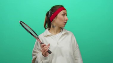 İzole edilmiş yeşil arka plan önünde tenis raketi tutan kadın beyaz kazak giyiyor ve kırmızı saç bandı takıyor. Spor ortamında odaklanma ve kararlılık ifadeleri sergiliyor.